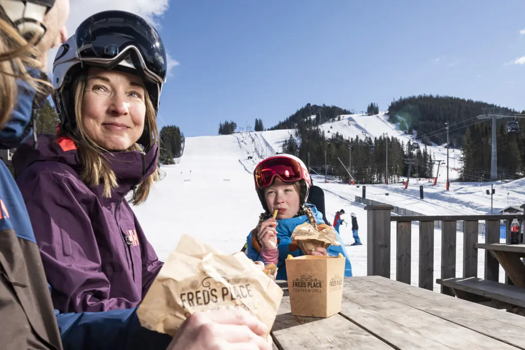 En familj, mamma, pappa och dotter, sitter på Fred´s uteseservering och äter hamburgare i solen.