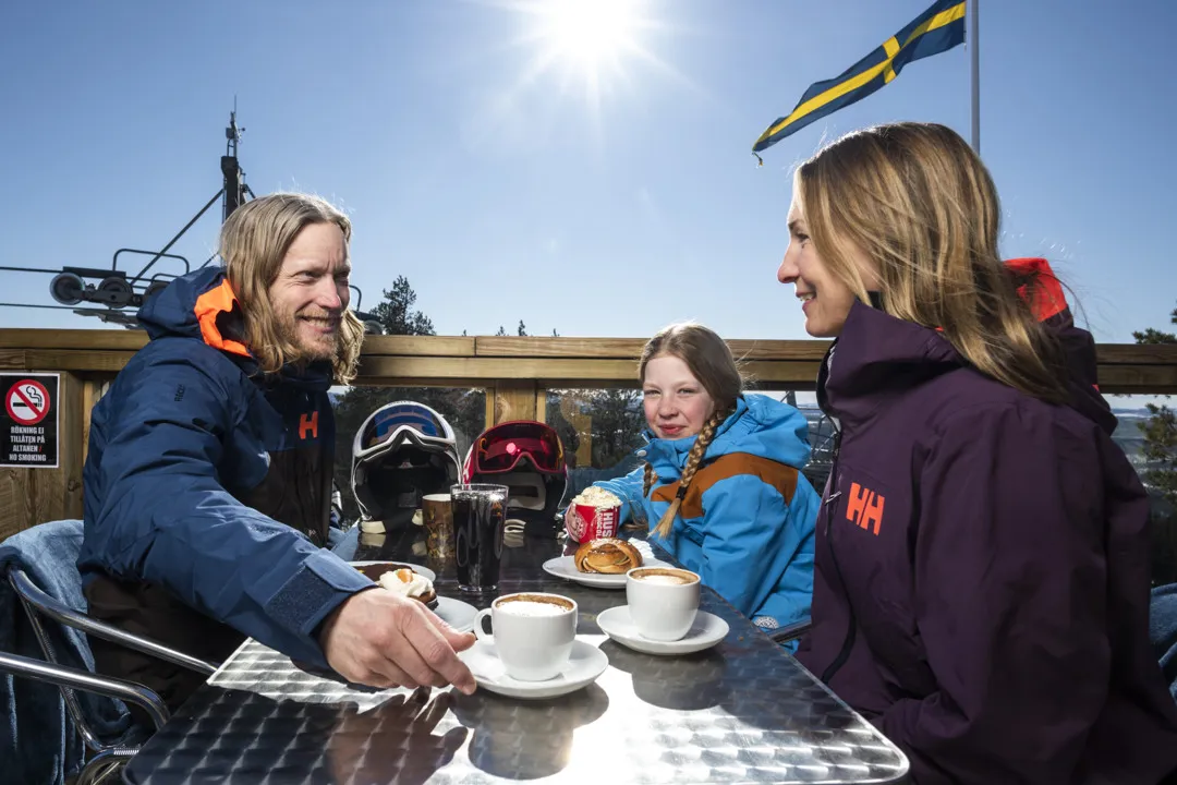 En familj sitter på Toppstugans uteservering och fikar. Det är blå himmel och sol.