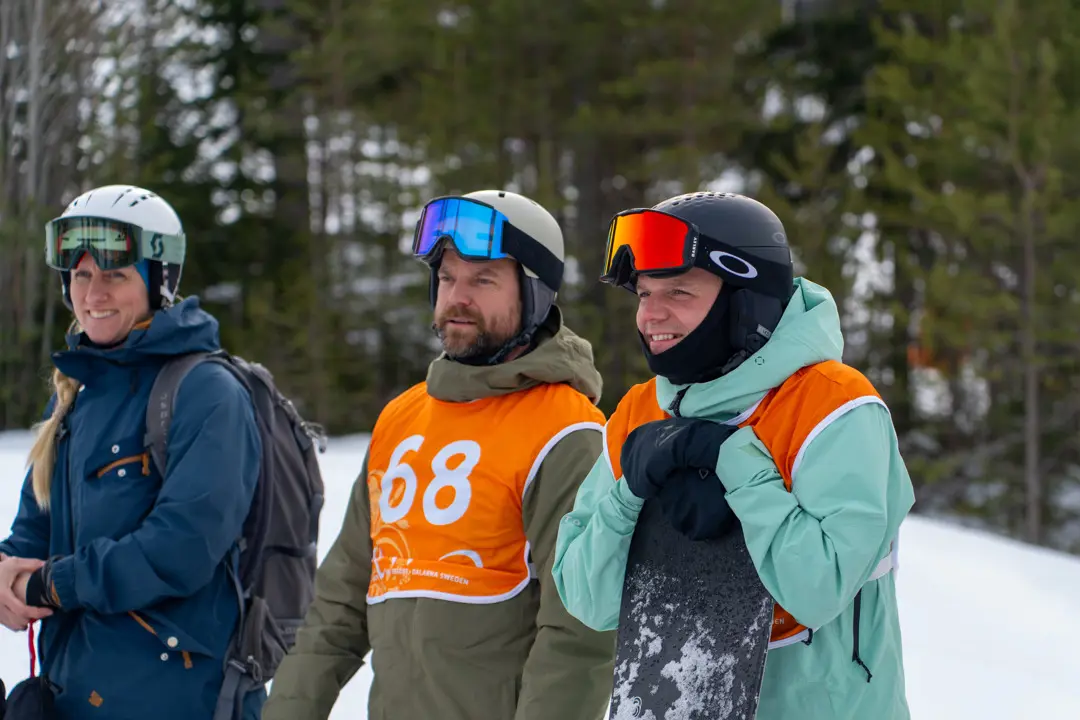 Two exhilarated gentlemen with bib numbers on their chests and a lady are watching as the competitors race down Ormtungan.